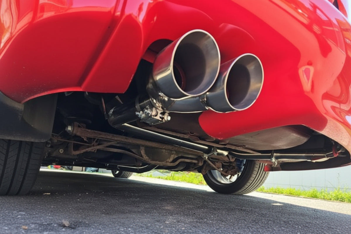 A close-up low-angle shot of a red car's dual chrome exhaust pipes mounted beneath the rear bumper, with asphalt pavement and a sliver of grass visible in the background.
