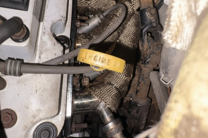 A close-up view of a used car engine bay, highlighting a bright yellow plastic dipstick filled with yellow engine oil, surrounded by metal parts, hoses, and protective wrap.