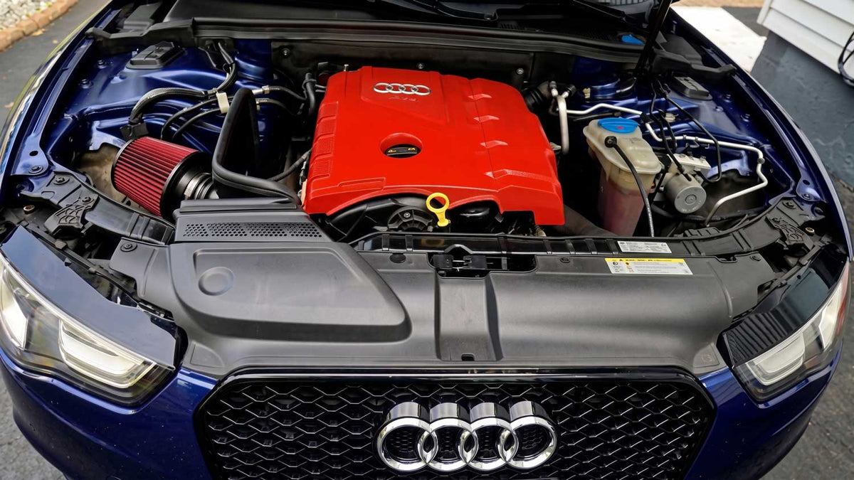 Close-up of the engine bay of a blue Audi car with the hood open, showing a bright red engine cover and red air filter, with the front grille and Audi logo visible.