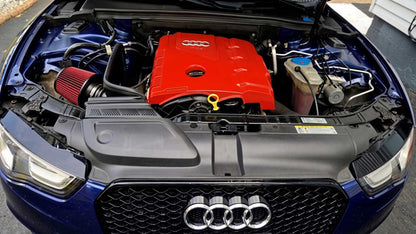 Close-up of the engine bay of a blue Audi car with the hood open, showing a bright red engine cover and red air filter, with the front grille and Audi logo visible.